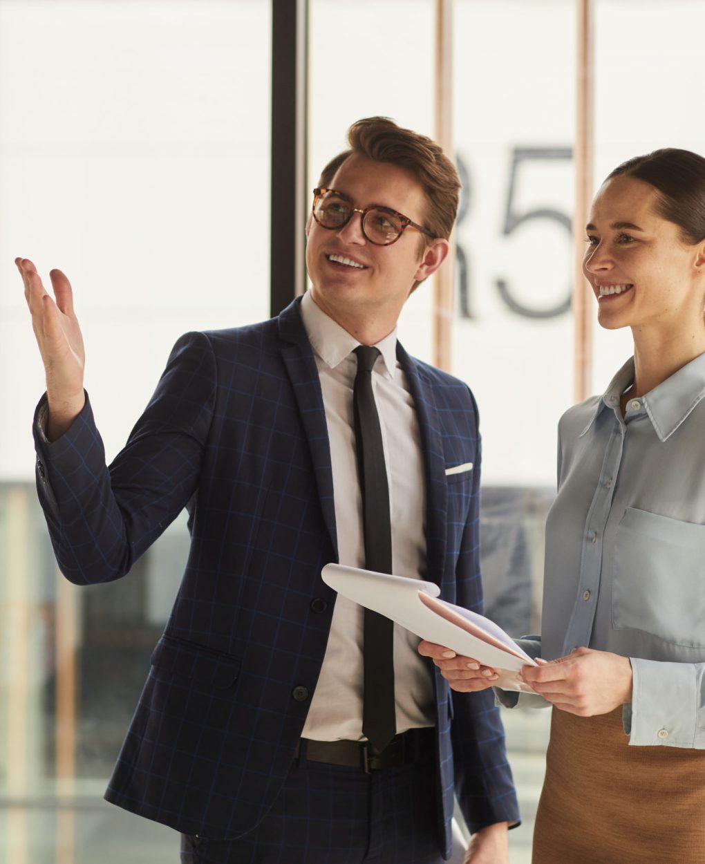 Waist up portrait of smiling real estate agent discussing property with female client and pointing up while standing in empty office building interior lit by sunlight, copy space