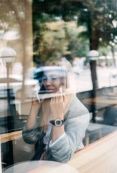 Self-employed young woman working for oneself as freelance or owner of business. Young freelancer woman working with laptop and talking by cellphone in cafe, view through the window.